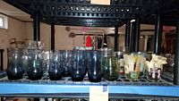 Wide view of assorted glasses on a black rack showing 9 clear plain glasses, 2 greenish tinted glasses, 4 floral pattern sour cream glasses, and other glassware.