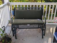 Full front view of the metal patio glider showing the green cushions and metal mesh seating and backrest. Glider placed on concrete porch with white railing in the background.