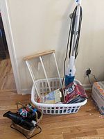 Photo showing white Easy Home stick vacuum leaning against wall beside white plastic laundry basket filled with various items including drying rack, microfiber cloths, and boxed iron.