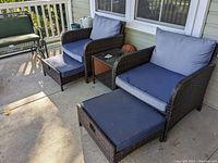 Two dark brown woven outdoor chairs with blue cushions and matching pull-out footstools, positioned on porch. Side table with glass top between them. Visible dirt and wear on cushions and furniture.