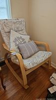 Side angled view of the Ikea slated wood rocking chair with floral cushion and throw pillows, showing wooden curved rockers and hardwood floor.