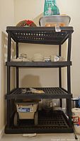 Front view of 4-tier black plastic shelving unit with various household items on shelves including dishes and containers. Unit shows signs of dust and needs cleaning.