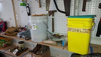 Overview of white bucket, yellow brush container, boxes with hardware and tools on a shelf against peg board.