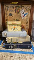 Photo showing wooden recipe box labeled 'Recipes' with colorful floral decoration atop a stack of three vintage cookbooks including two 'Joy of Cooking' books and one 'The New York Times Cook Book' with the framed spice cabinet in the background.
