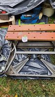 Folded work table showing wooden slats with holes and gray metal foldable legs, positioned on tarp on grass.
