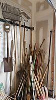 Wide view showing large assortment of yard tools including shovels, rakes, level, and other garden tools leaning against the wall and pegboard
