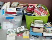 Photo showing two large plastic storage bins filled with stacks of assorted greeting cards and stationery items with various card designs visible.