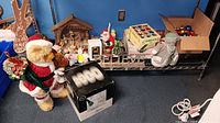 Wide shot of shelf with teddy bear Christmas plush, nativity scene, Santa figurine, and boxed glassware on floor.