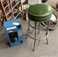 Metal stool with green worn upholstered seat and silver metal frame next to a blue coin operated payphone on carpeted floor