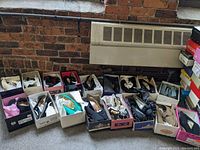 Wide view of multiple women's shoes in open shoeboxes arranged on floor against brick wall and heater.