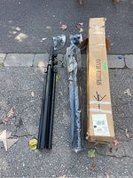 Two black speaker tripods and one cardboard shipping box labeled 'Speaker Stand' on asphalt surface, some packed components still in plastic wrapping.