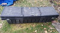 Full view of large black plastic truck bed tool box on grass, showing three lid sections, side push-button latches, and overall exterior shape and condition.