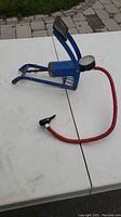 Side view of blue metal foot pedal air pump with attached pressure gauge, standing on a white surface outdoors.