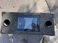 Front view of carpeted rectangular enclosure with two bass ports and central viewer window showing subwoofer cones inside.