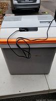 Front view of gray and white Mobicool portable cooler with orange handle, power cord attached on top.