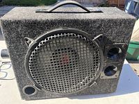 Front view showing speaker behind metal grille, circular ports, and carpeted enclosure.