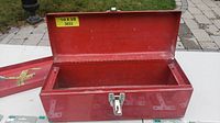 Empty red metal tool box with latch displayed open on outdoor surface.