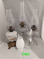Front view of all three oil lamps and three chimneys showing different glass designs and bases including footed milk glass and clear glass with square base.