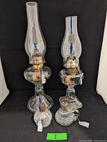 Four antique oil lamps including two tall glass lamps with chimneys, a smaller finger lamp and a laboratory burner, all with metal burners and wicks visible.