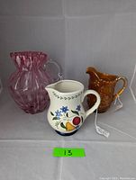 Three pitchers displayed against white backdrop: marigold carnival glass, cranberry swirl blown glass with crack, and ceramic with painted flowers and fruit.