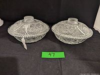 Pair of clear glass covered dishes with embossed hearts and geometric patterns, both with lids and square-shaped handles, displayed against a black background.