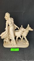 Front view of white vintage sculpture showing lady with two large dogs and a puppy at her feet on an oval base.
