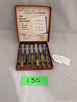 Six vintage Dinkee spreader knives with Bakelite handles arranged in original red box. The inner lid of the box features printed advertising text describing the use as an ideal spreader for butter, cheese, jam, and a perfect fruit knife.
