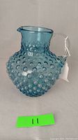 Front view of a vintage blue hobnail glass pitcher with clear handle and a tag attached. No chips or cracks visible.