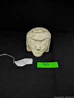 Front angled view of a vintage head sculpture showing stylized facial features and geometric top design on a black background with a green tag and price label.