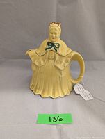 Front view of vintage ceramic teapot shaped like an old lady wearing a bonnet and shawl, tagged with number 136.