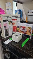 Two Hamilton Beach single serve blenders in original boxes - black and red models, plus MainStays digital kitchen scale in box and a white analog kitchen scale on counter.
