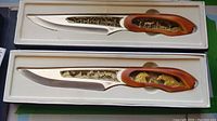 Two decorative collectible knives in display boxes, showing entire blades and handles with gold-colored carvings of wolves and landscapes.