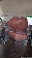 Two Wilson NFL Soft Grip footballs stacked vertically in clear acrylic display cases showing signatures on sides of each ball.
