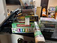 Lots of new and unopened board games stacked, including Deal Or No Deal, Monopoly, Life, with packing paper visible and some DVDs in the background.