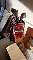 Red and white Callaway golf bag with clubs inside, surrounded by various boxes and items, showing the side and strap of the bag.
