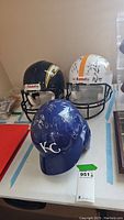 Three helmets displayed on a table: blue KC Royals baseball helmet, black Chargers football helmet, white signed football helmet.