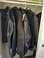 Photo showing five black and dark brown men's coats hanging side-by-side, displaying variety of styles including leather, insulated, and casual fabric jackets.