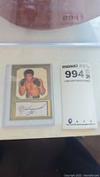 Photo showing Muhammad Ali trading card in protective plastic sleeve alongside auction notes.