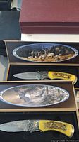 Photo of two pocket knives in boxes featuring detailed wildlife designs and natural scenes inside box lids.