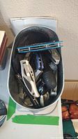 Top-down view of metal bucket filled with various pocket knives and cutters, showing a variety of handle designs and folding mechanisms.