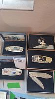 Photo showing five pocket cutters in boxes, four have decorative handles with rural and wildlife scenes, one is a folding metal cutter with two blades.