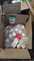 Cardboard box filled with multiple golf balls, some individually wrapped in plastic bags alongside a TaylorMade ball pack.