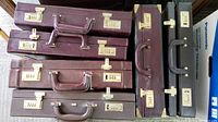 Stacked view of 11 hard leather attache cases in burgundy hues showing combination lock clasps, handles and hard edges.