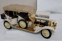 Front left angle view showing open hood with detailed gold brass engine, white and gold body, beige fabric roof, black interior, and spare tire on side.