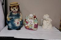 Full view of the lot showing the dog cookie jar, Buddha figure with children, and white Geisha incense burner figurine on a white cloth.