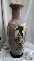 Side view of the large ceramic vase showing detailed hand-painted scenes on a pink background with floral patterns. Vase mounted on a wooden stand.