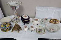Wide shot of eleven porcelain ring and bon bon boxes arranged on a table, including a cherub candlestick holder, a blue and gold floral box, and a gold metal turtle-shaped box.