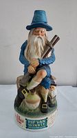 Front view of Rip Van Winkle porcelain bottle in blue attire holding a rifle, sitting on a log with a ceramic jug on base