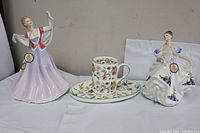 Wide view showing two Royal Doulton figurines and a floral china cup and plate set on a flat surface.