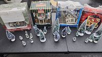 Four boxed Christmas village houses and multiple flocked trees displayed on table.